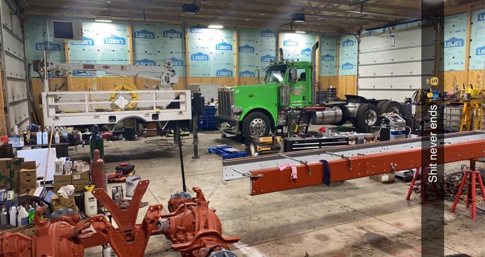 A mechanic's garage with a green semi-truck, a white truck bed, and tools; orange parts in the foreground.