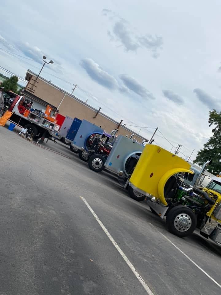 Semi-trucks parked in a lot, some with hoods open for repair. Cloudy sky overhead.