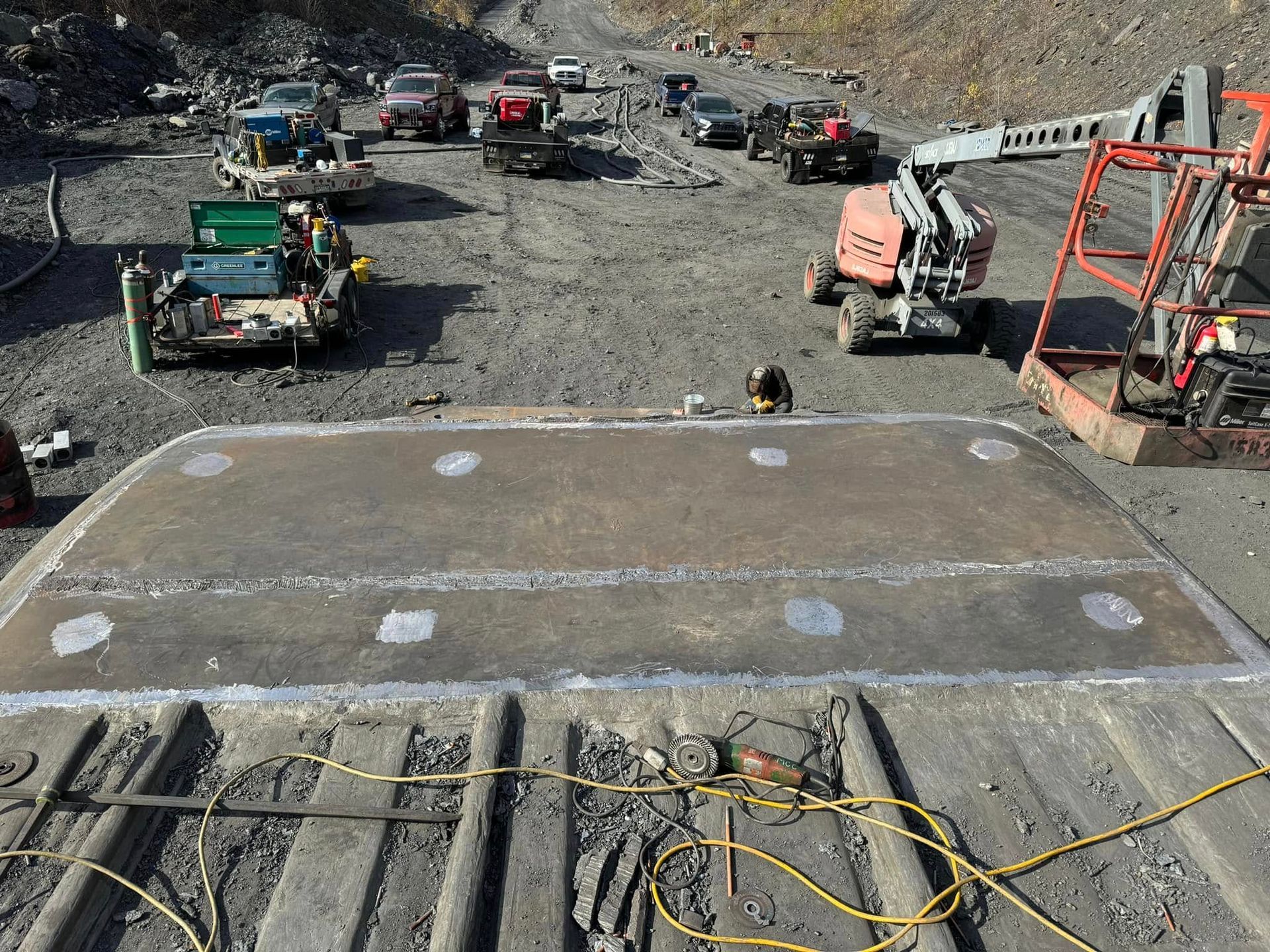 Large metal plate being welded in a rocky industrial setting, surrounded by vehicles and equipment.