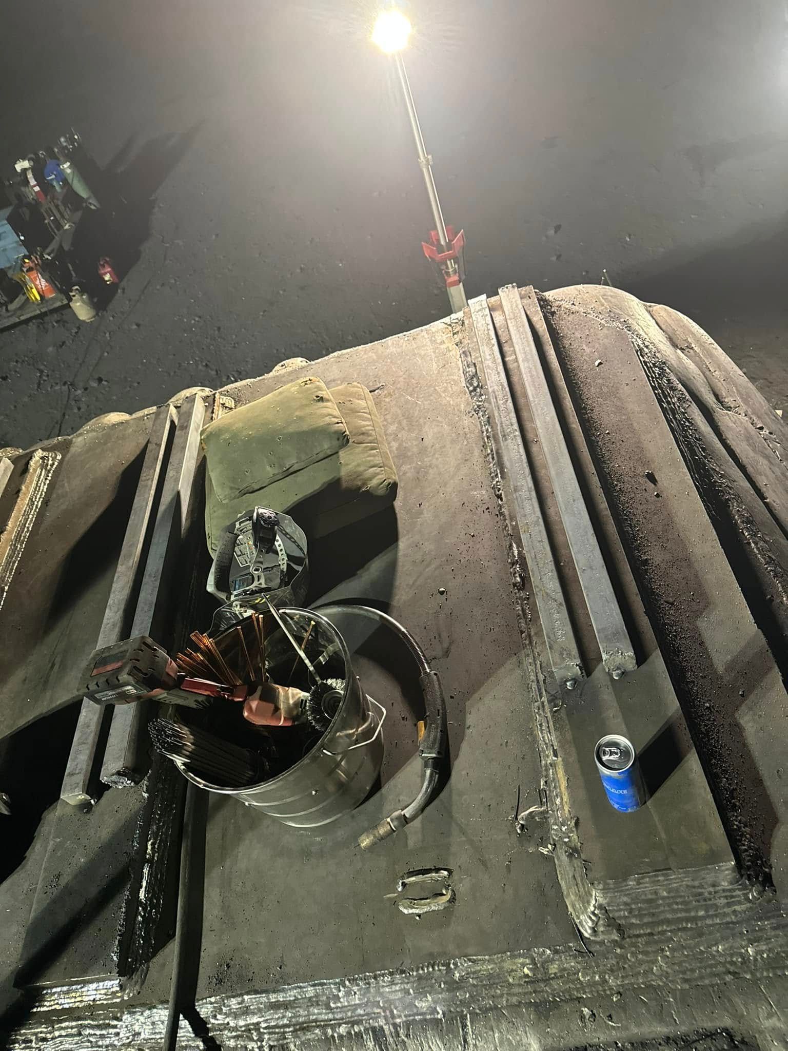 A camera on a tripod is attached to a large, dark, industrial structure, lit by a bright overhead light.