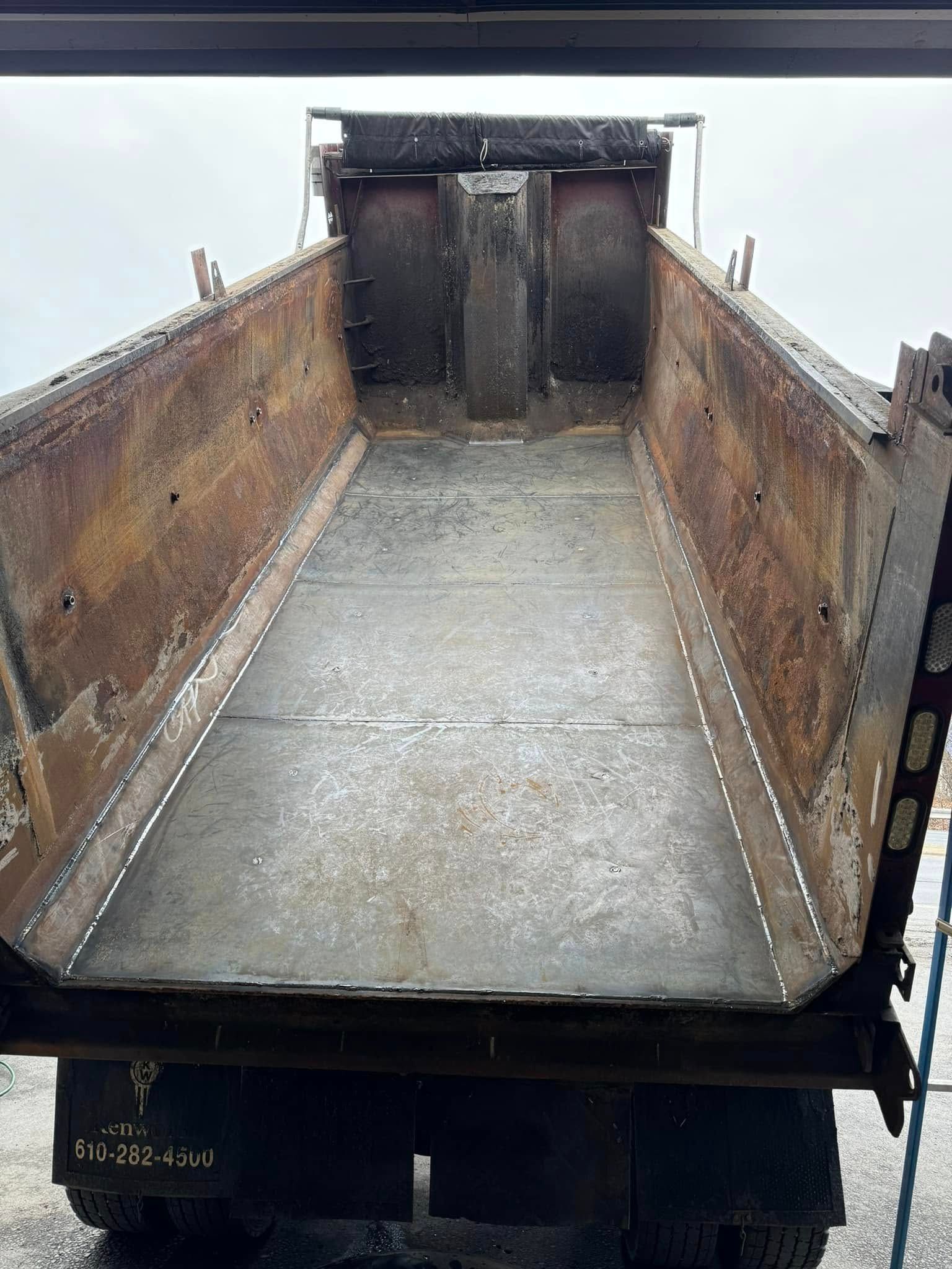 Empty, rusty dump truck bed, angled walls. White dotted lines mark repairs, overcast sky.
