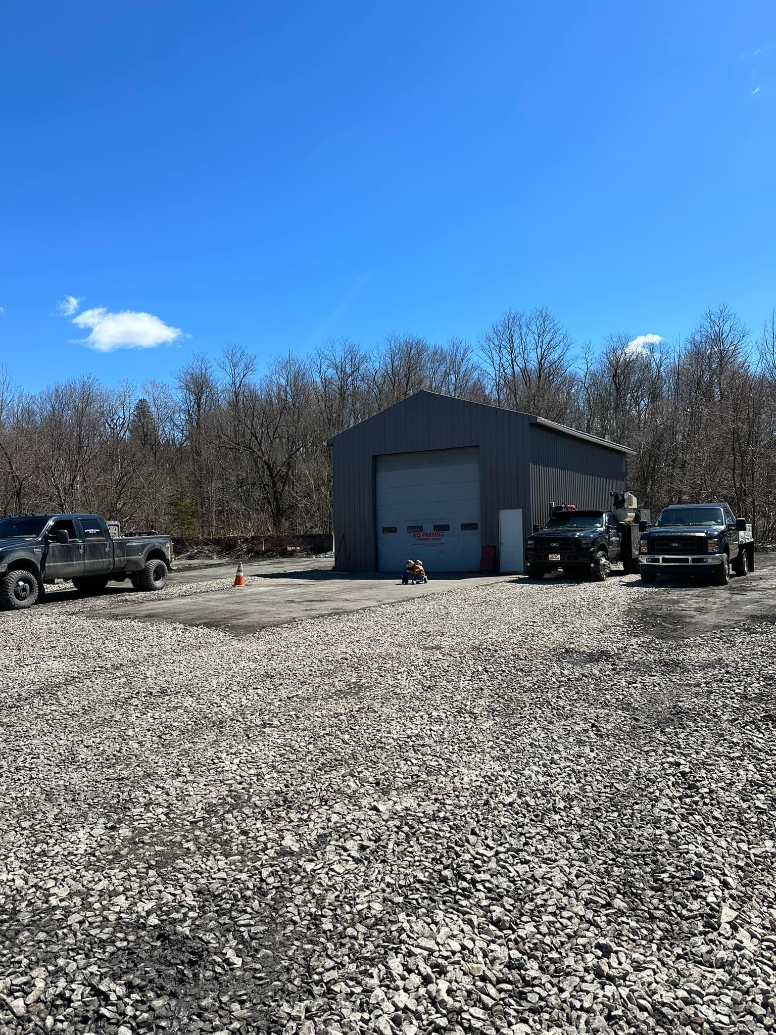 A gray metal workshop with garage door, surrounded by gravel. Trucks parked outside under a blue sky.
