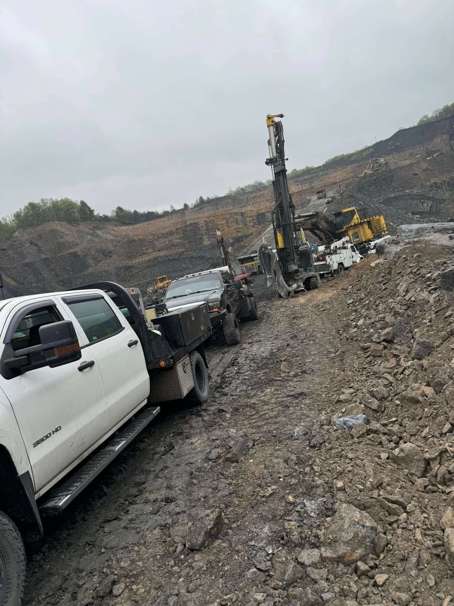 White truck towing drill rig on a rocky, coal-like path near a hillside on an overcast day.