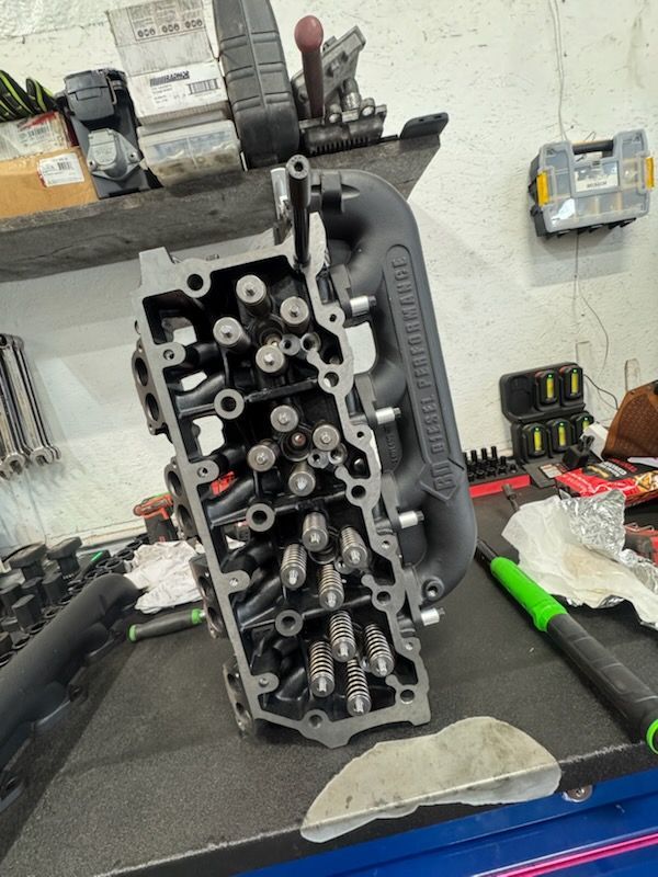 Engine cylinder head on a workbench, open with valves visible. Black intake manifold, tools in background.
