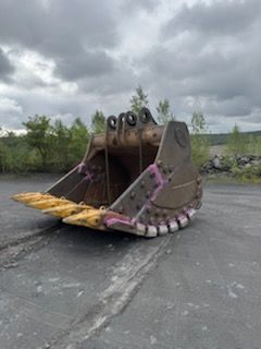 Large, worn metal excavator bucket on a gray surface, in an outdoor setting with a cloudy sky.