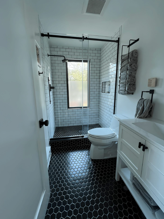 A bathroom features a walk-in shower with white vertical tiles, a glass door, black hardware, and a stone pebble floor.