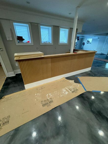 A newly installed wooden slat bar counter with white trim stands in a room with a glossy gray floor and three windows.