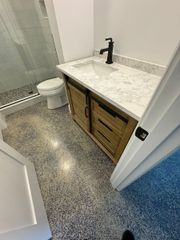 A modern bathroom with a wooden vanity, white stone countertop, black faucet, and speckled gray floor tiles.