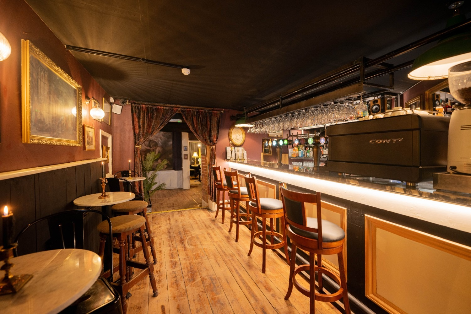 Interior of a dimly lit bar with a long counter and stools. Dark walls, gold-framed art, and wooden floors.