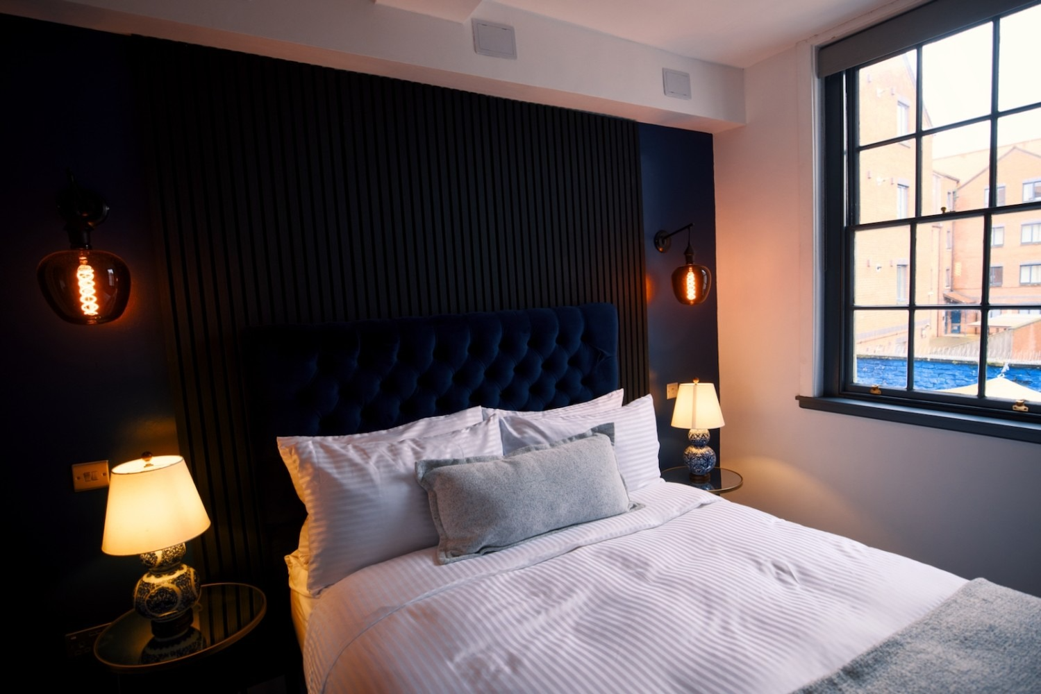 Bedroom with a tufted headboard, side lamps, and a window with a view of a brick building.