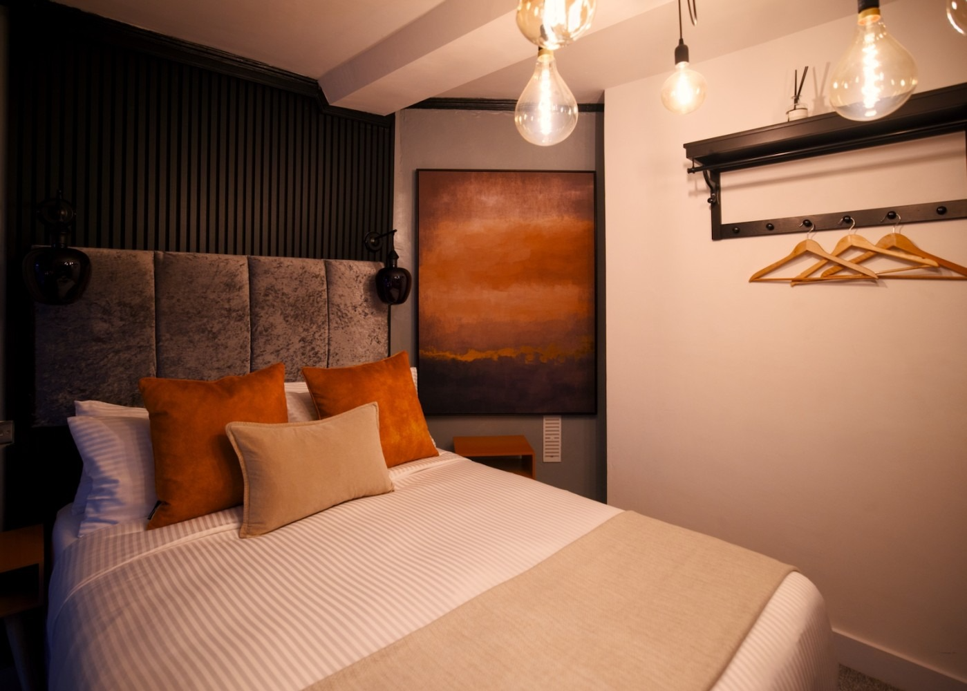 Cozy bedroom with bed, orange pillows, and abstract art, lit by hanging bulbs.