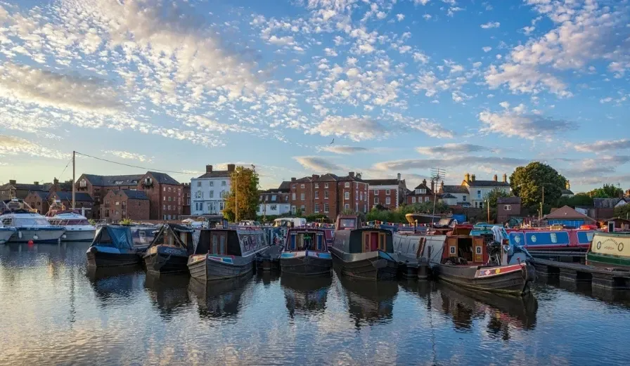 Narrowboats moored in a harbor, reflecting in the water, with buildings and a cloudy blue sky in the background.