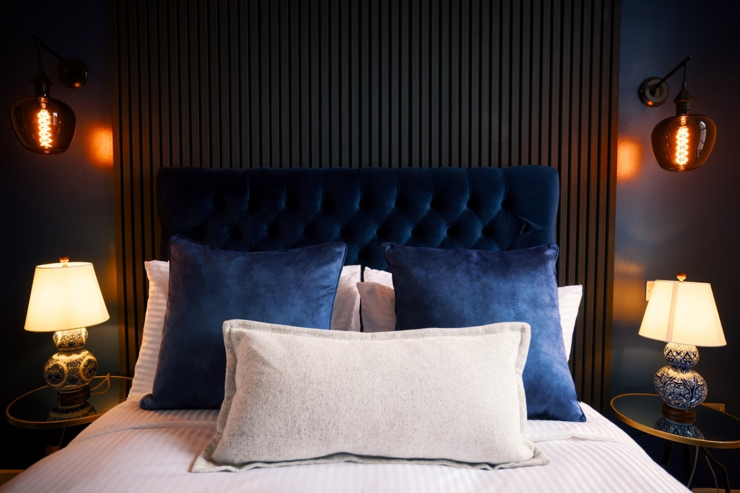Bedroom with blue tufted headboard, pillows, lamps, and dark wall paneling.