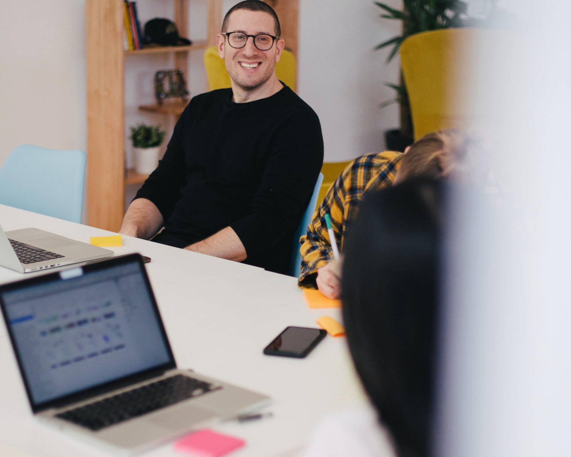 Man in black sweater smiles during a meeting at a table with laptops, a phone, and a person in a plaid shirt.