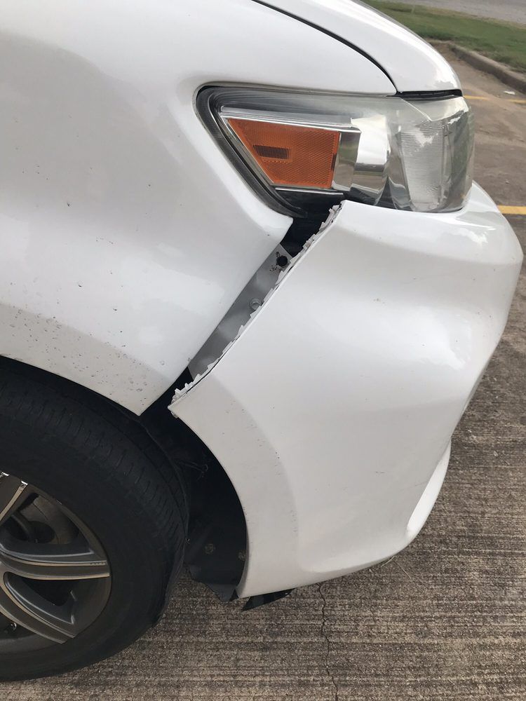 A white car with a broken bumper is parked on the side of the road.