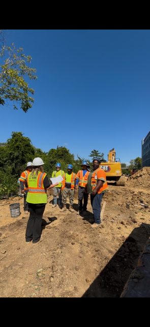 Construction workers in reflective vests and hard hats gather outdoors under a blue sky, near heavy machinery.