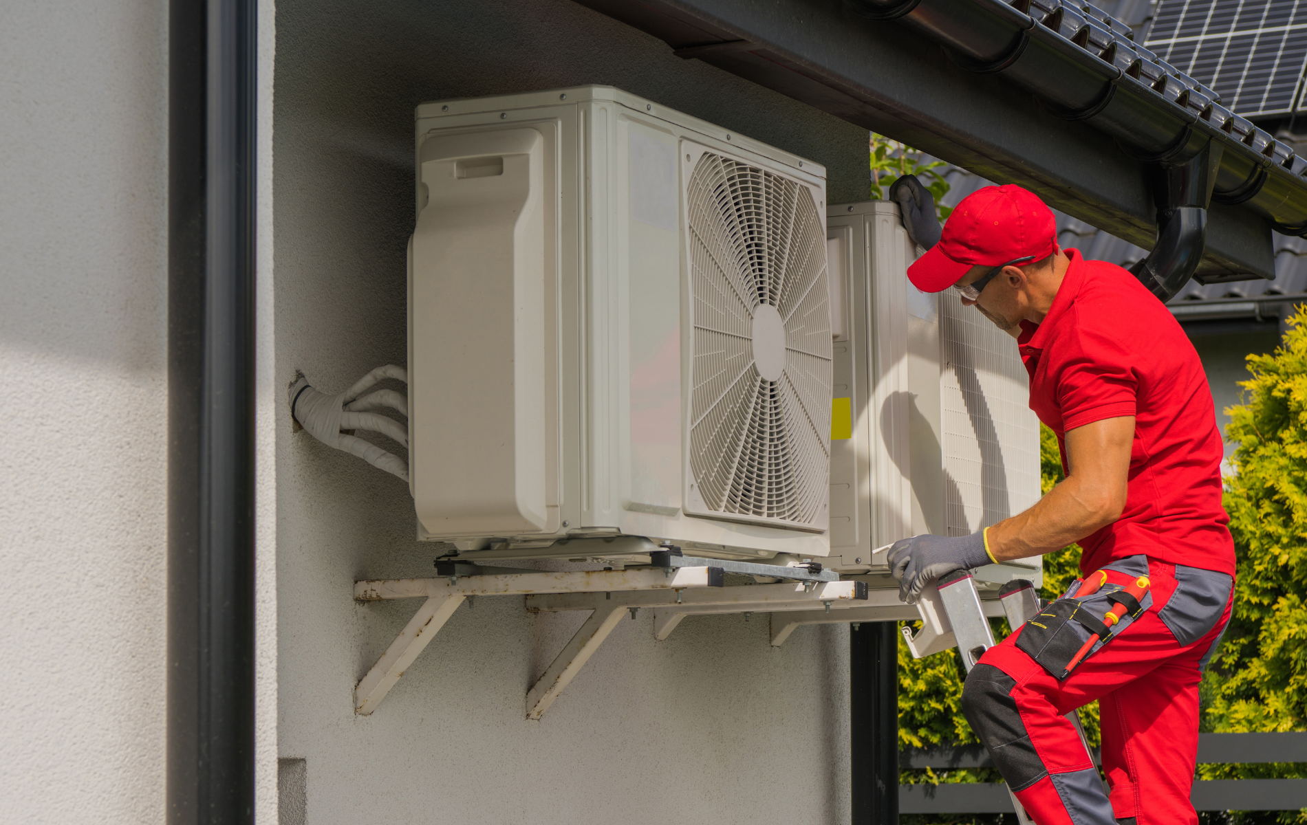 A worker in a red uniform and hat installs an air conditioning unit on a bracket outside a house.