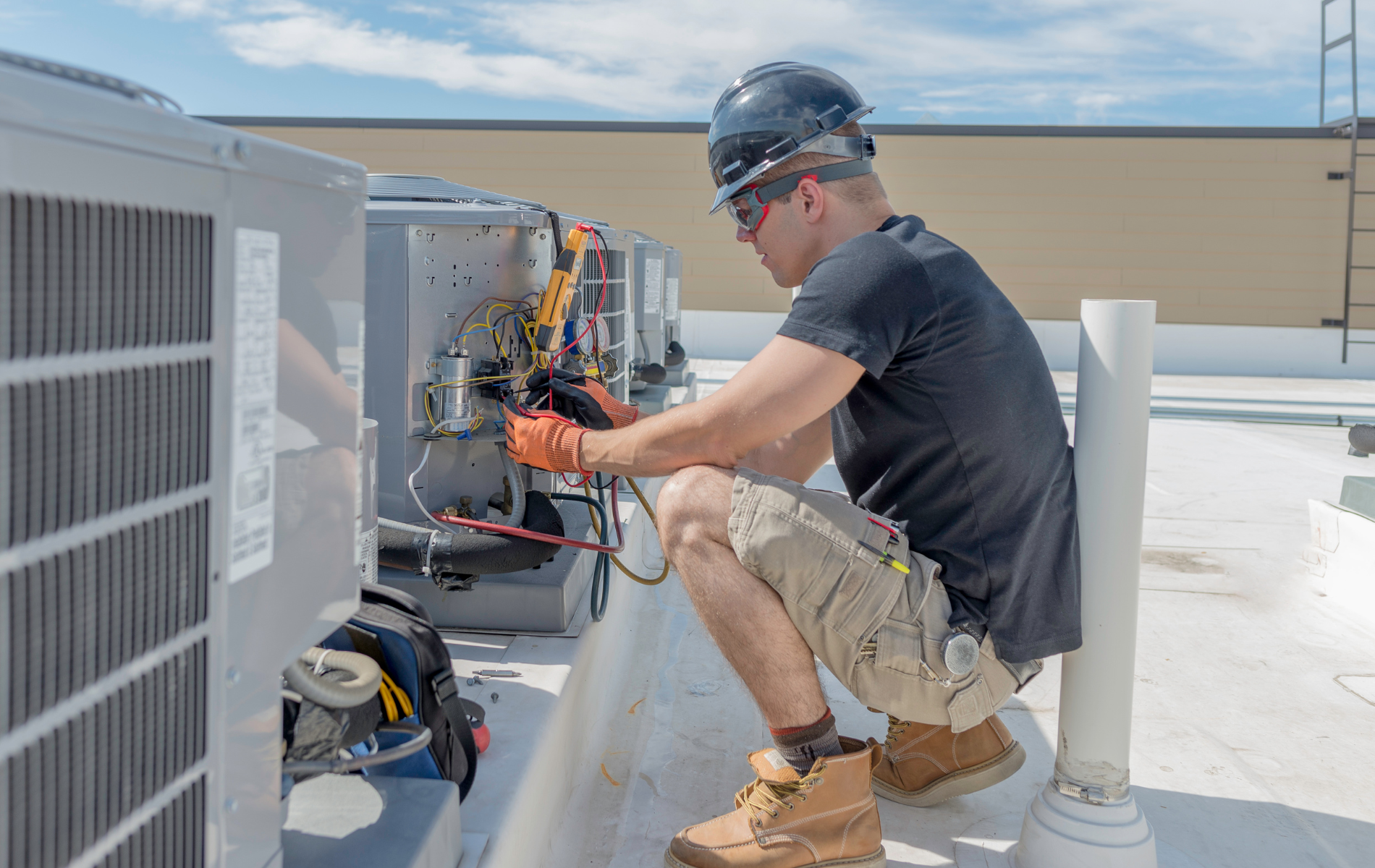 Technician kneels beside rooftop HVAC equipment, using tools on a bright sunny day.