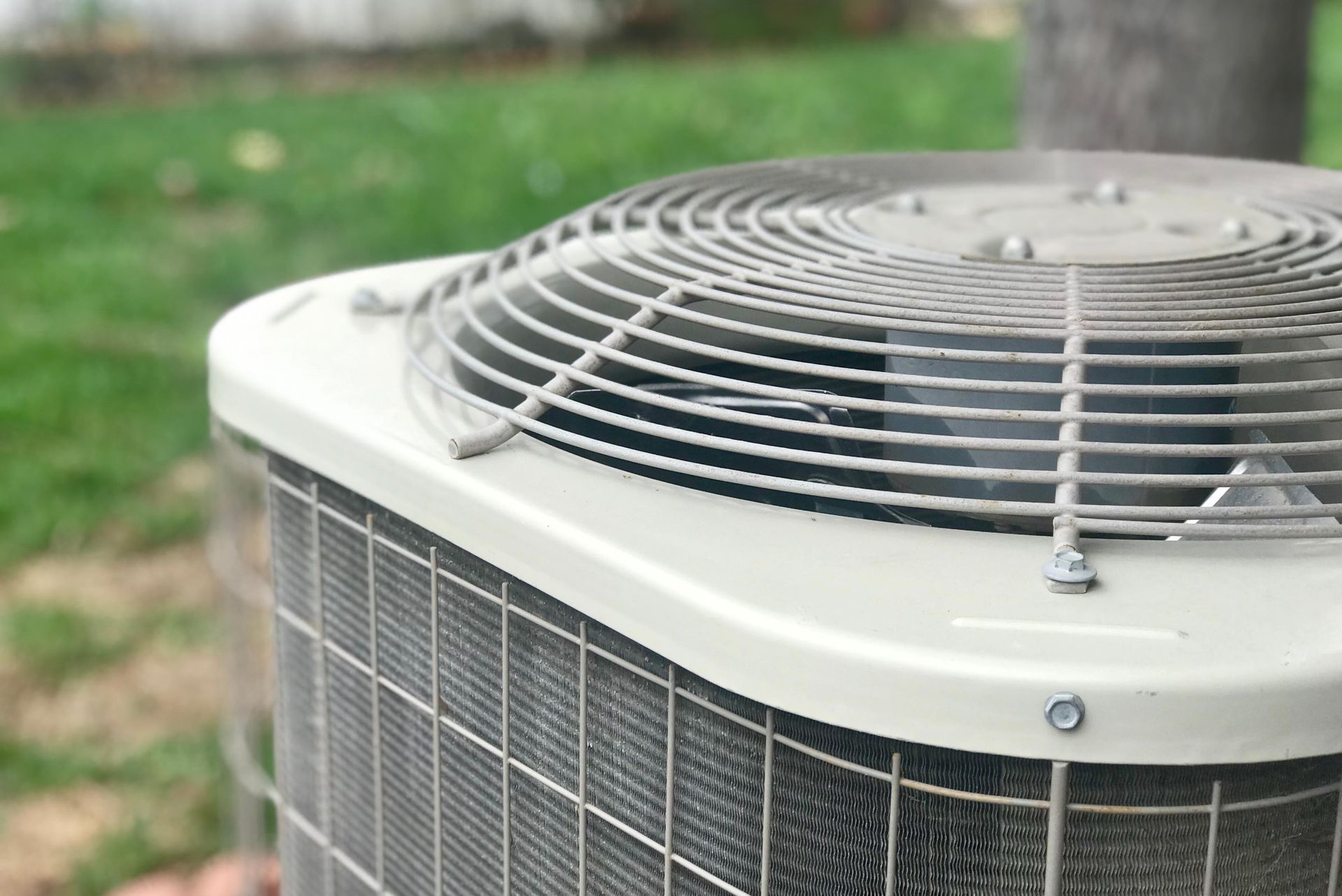 Air conditioning unit outdoors with metal grill and fan on a blurred green background.