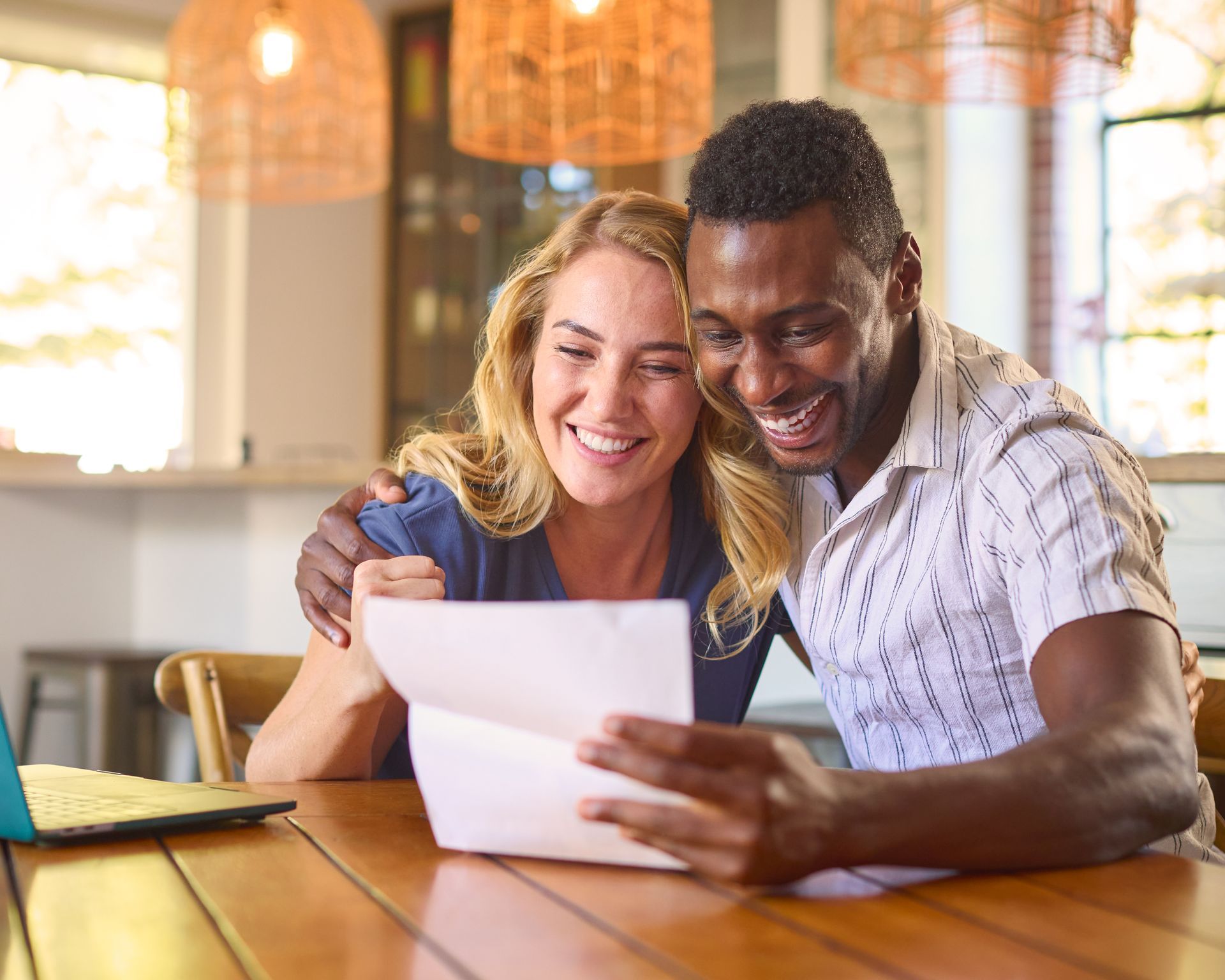 Couple smiling, looking at a document at a table indoors. One has arm around the other.