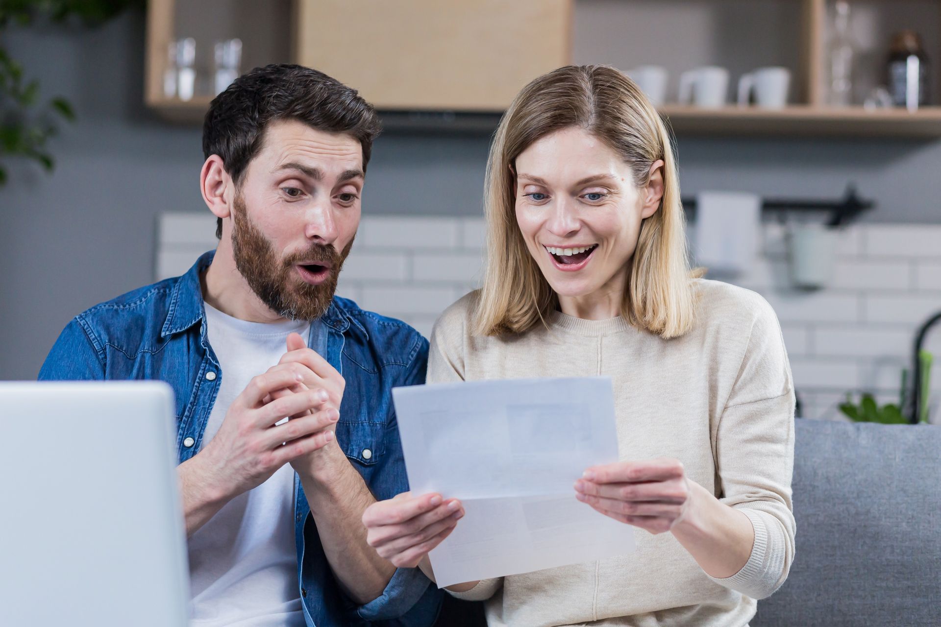 Two people smiling and reading a document at a laptop in a bright home kitchen