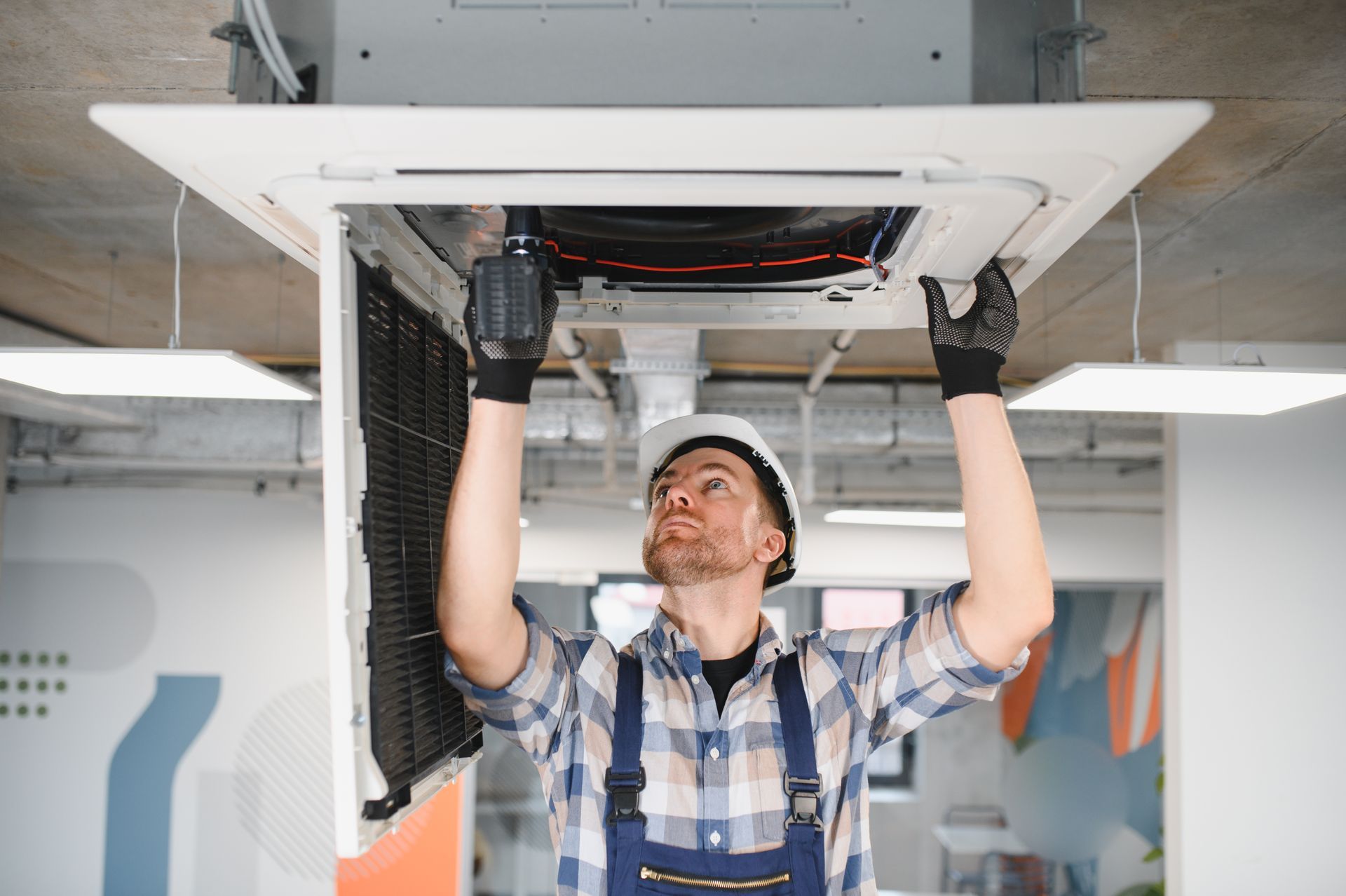 A technician in overalls and a hard hat repairs an overhead air conditioning unit.