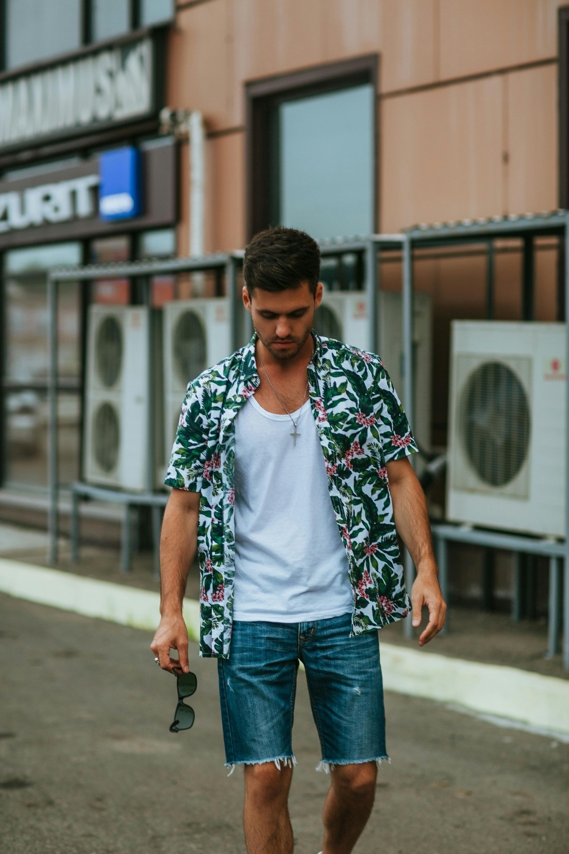 Man in floral shirt, white tank, and denim shorts walks outdoors, holding sunglasses.