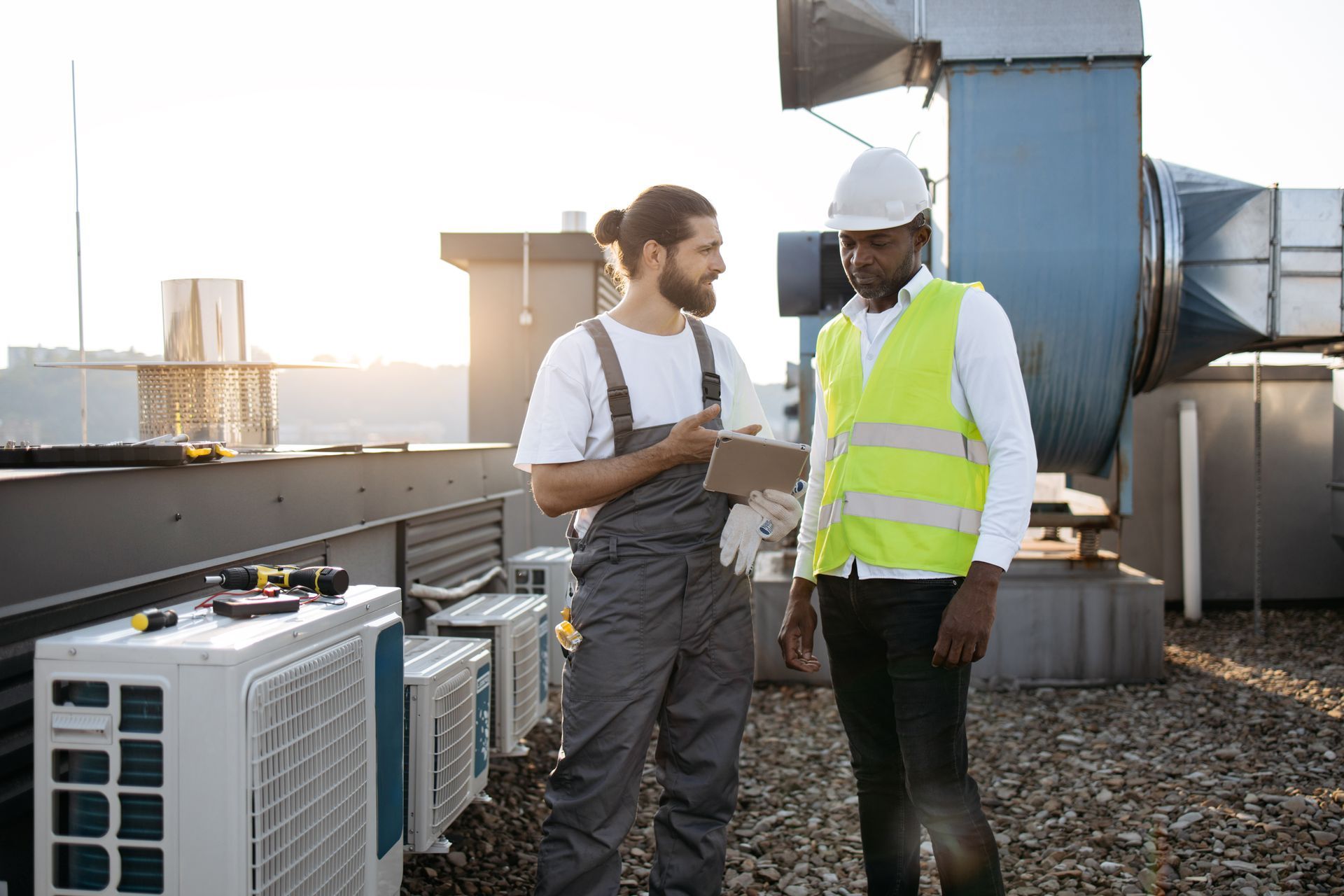 Two technicians on a rooftop discussing plans near HVAC equipment. One holds a tablet.