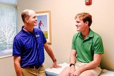 A healthcare provider in blue shirt converses with a patient in a green shirt in an exam room.