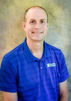 Man wearing a blue polo shirt smiles; a logo is on the shirt. Background is a neutral, mottled color.