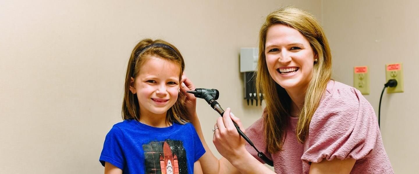 A healthcare worker examines a child's ear with an otoscope in an exam room. Both smile.