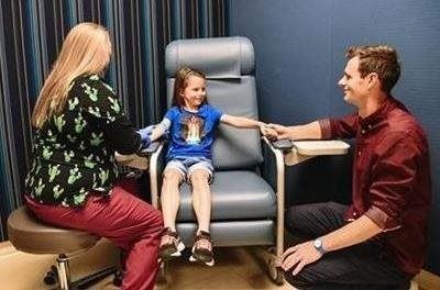 A young child sits in a medical chair. A person in scrubs and a person in a button-up shirt attend to the child. Blue walls.
