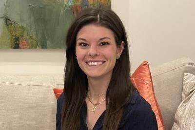 Woman smiling, sitting on a couch, wearing a blue shirt and necklace; painting in background.