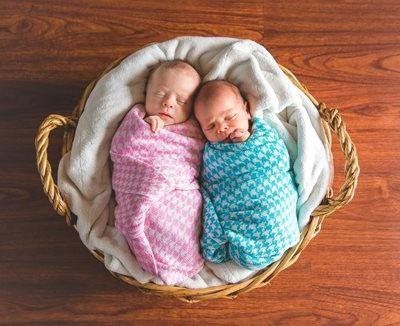 Two swaddled babies sleeping in a wicker basket on a wood surface; one in pink, one in turquoise.