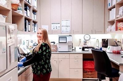 Woman in scrubs smiles in a medical lab. She wears gloves, is surrounded by equipment and supplies.