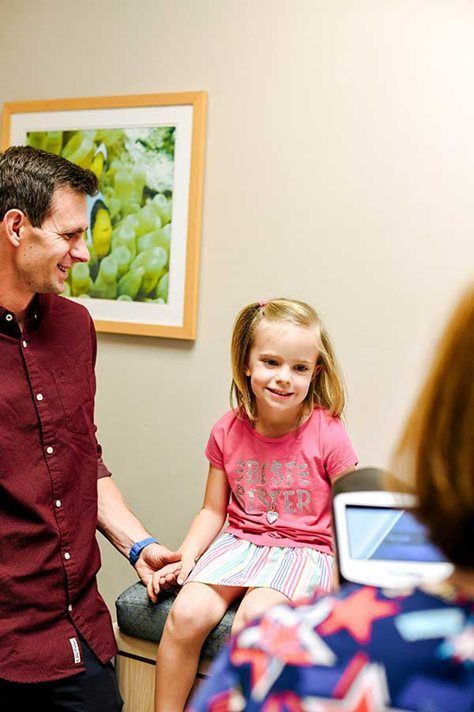 A father and daughter at a doctor's visit, smiling. They sit with a medical professional in an office setting.