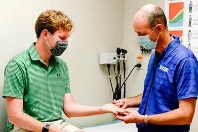 A doctor examining a patient's hand in an examination room; both are masked.