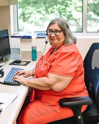 Woman in orange scrubs at a desk wearing a headset. She is typing at a keyboard, smiling, in an office with a window.