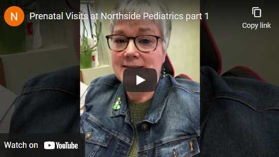 Woman in denim jacket, glasses, green pin, recording video.