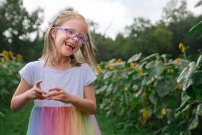 Girl in glasses laughs in a field of sunflowers, wearing a rainbow dress.