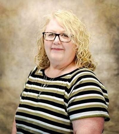Woman with curly blonde hair and glasses, wearing a striped shirt, smiling in a studio setting.