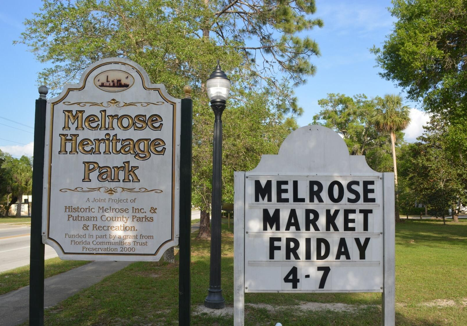 Sign for Melrose Heritage Park and market, Friday 4-7. White signs with black text, a lamp post, and green grass.
