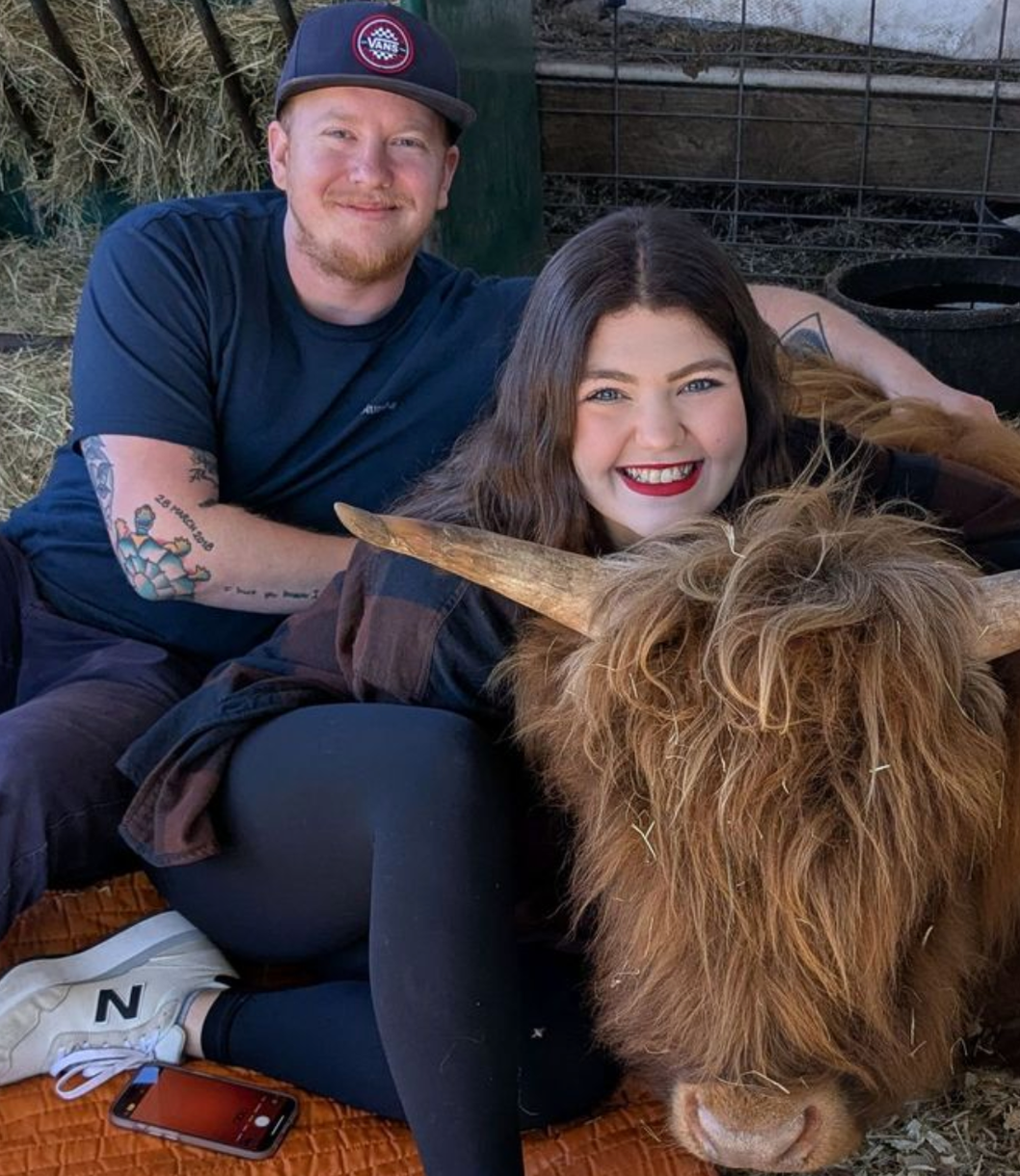 Couple smiles next to brown Highland cow with long fur in a Florida animal farm