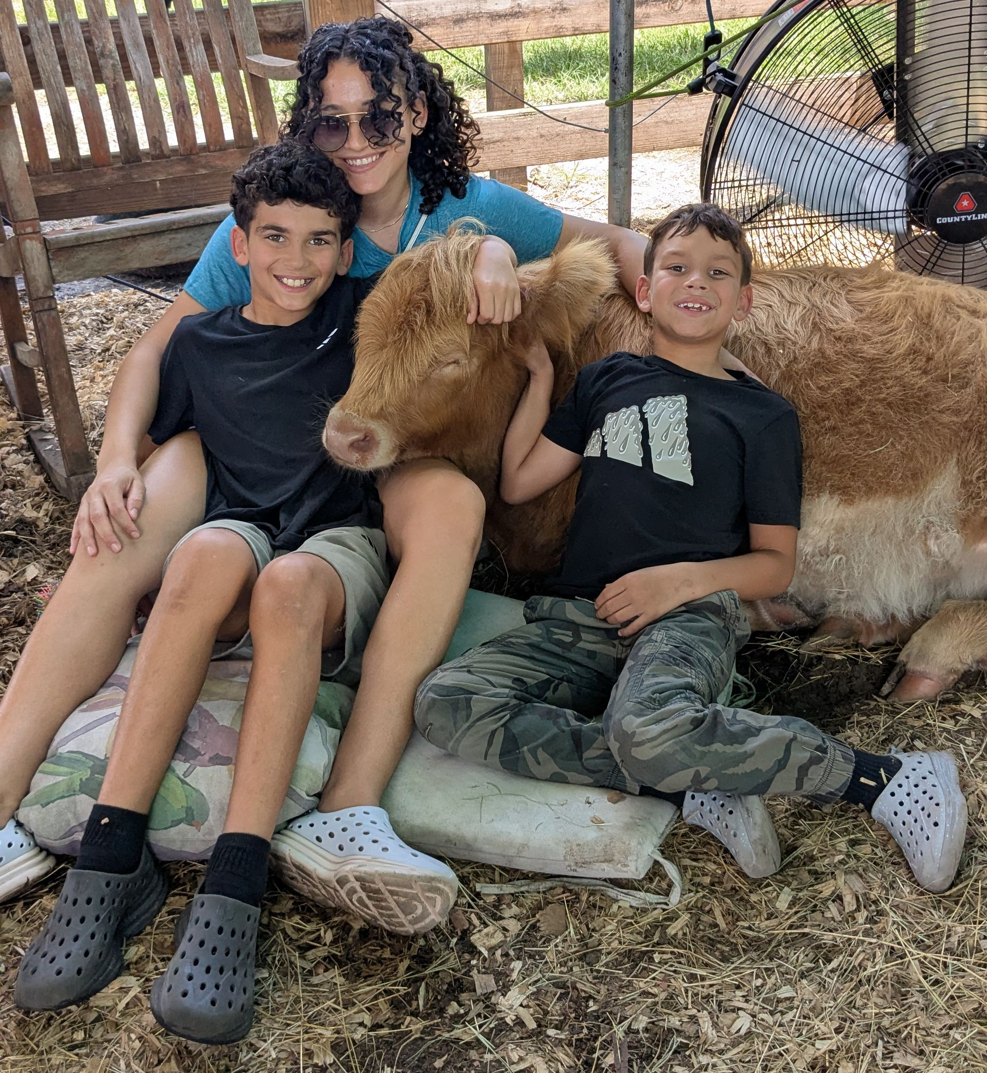 Three people sit with a light-brown cow. Two boys smile while a person with curly hair smiles in a Florida animal farm