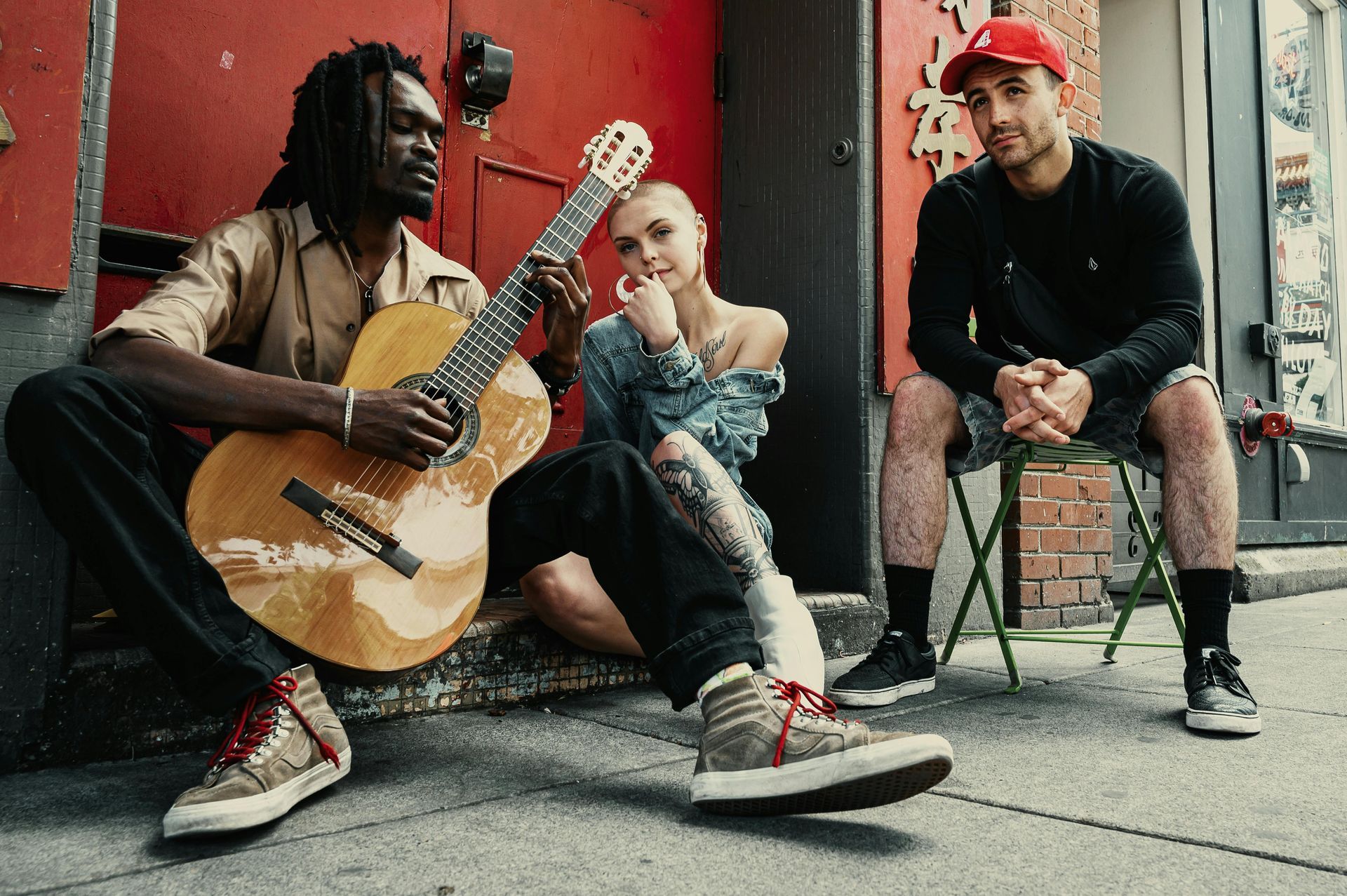 Three people on a city sidewalk. One plays guitar, another sits, and the third wears a red hat.