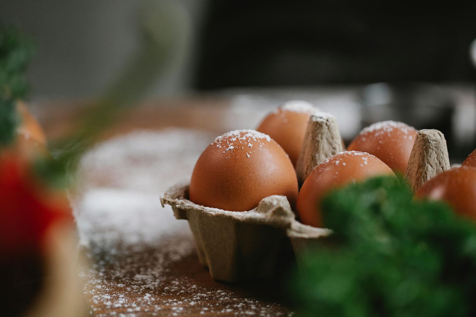 Brown eggs in a carton on a wooden surface dusted with flour, next to green leafy vegetables.