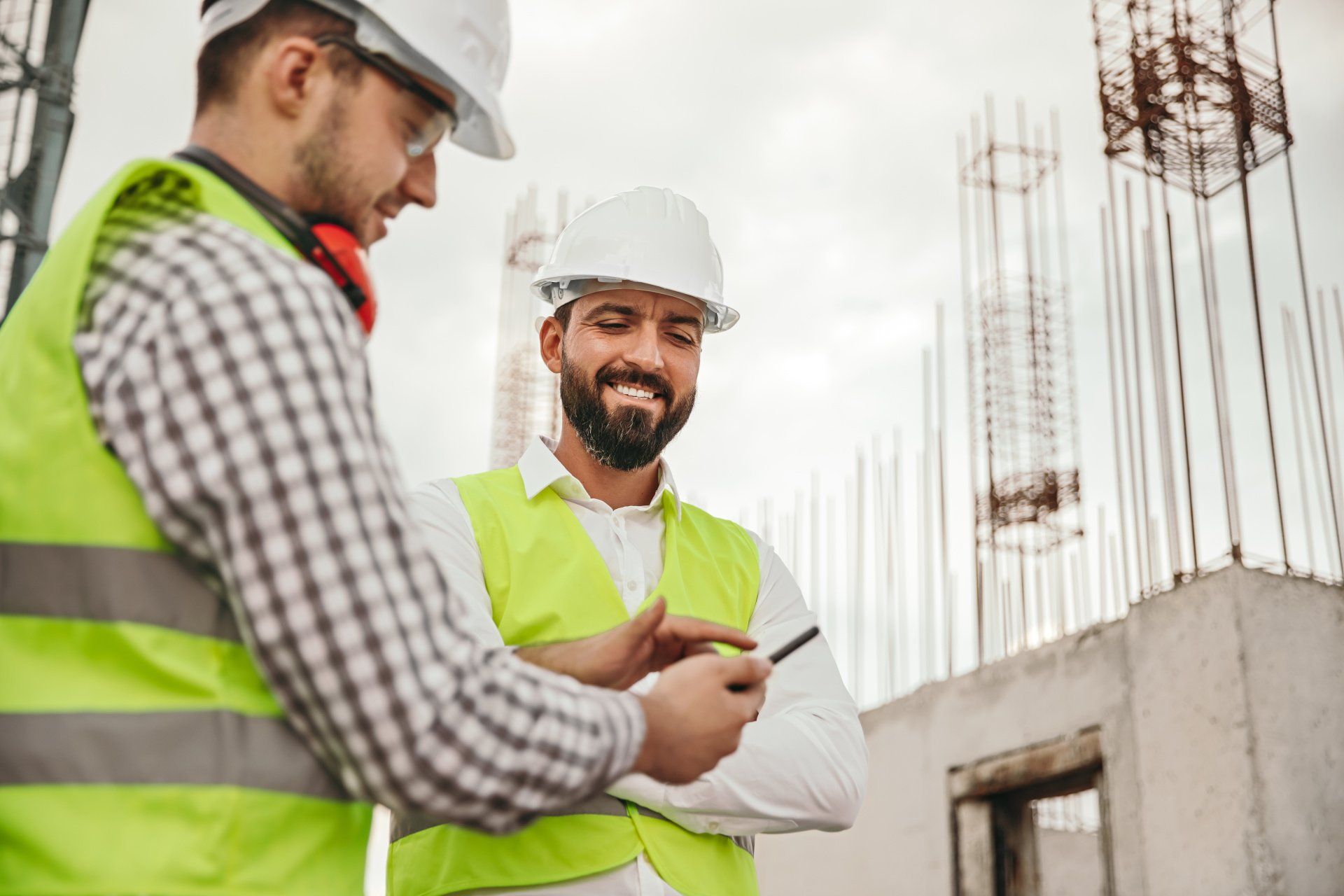 Two construction workers wearing hard hats and safety vests looking at a phone on a construction site.