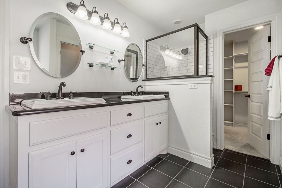 Modern white bathroom with black countertops, black floor tiles, and a walk-in shower.  Double sinks with oval mirrors and a walk-in closet.