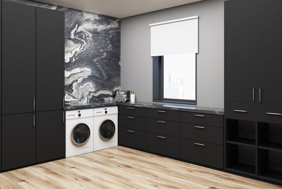 Modern laundry room with black cabinets, marble backsplash, and white appliances. Wooden floor and a window with a white shade.