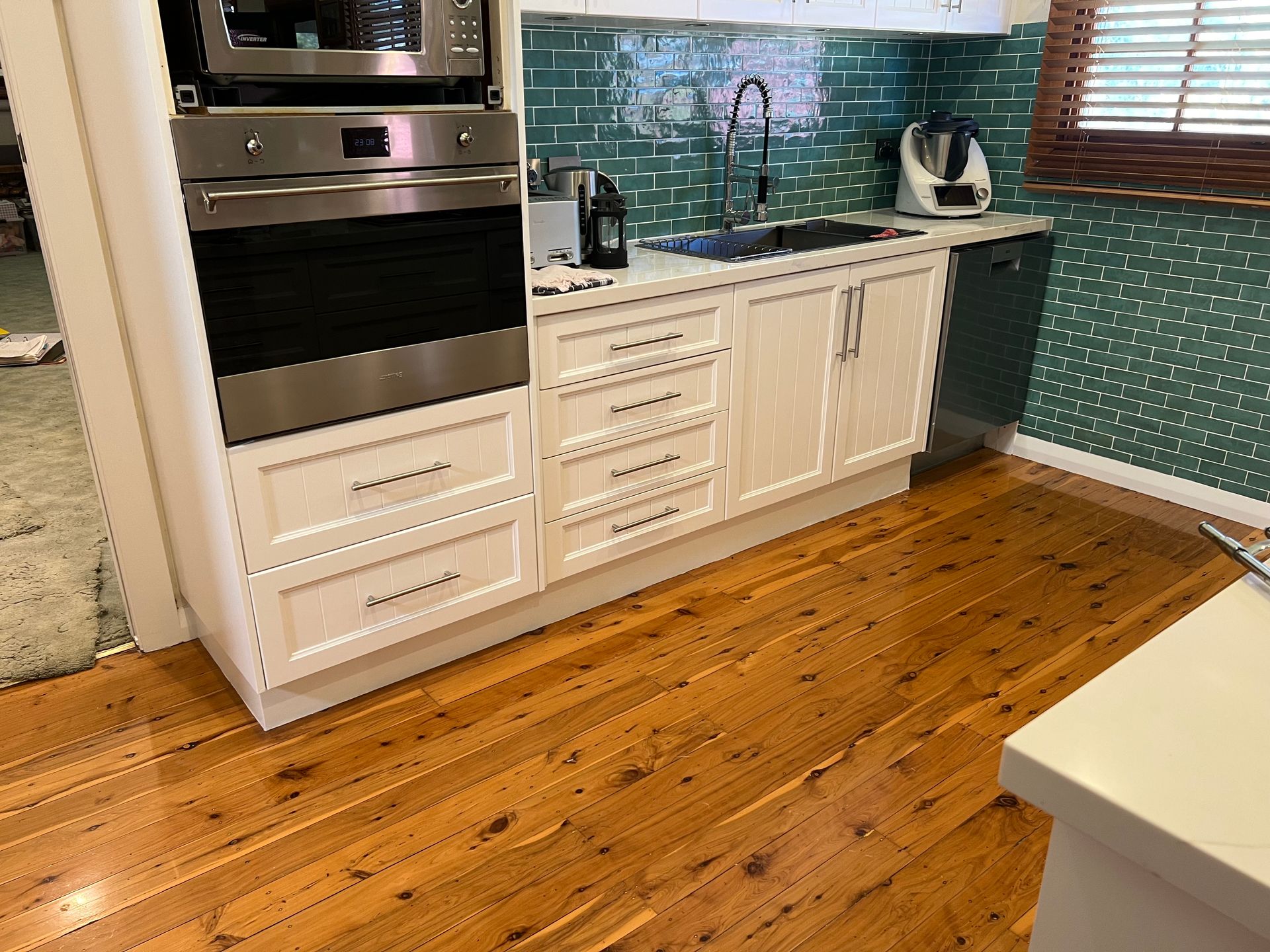 Kitchen with white cabinets, stainless steel appliances, and wooden floors. Teal backsplash and brown blinds.